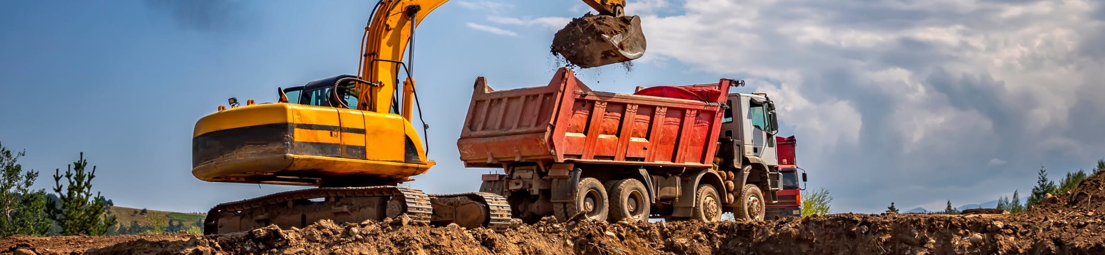 Dirt being lifted into a truck in an open space with sky and some trees visible in the background 