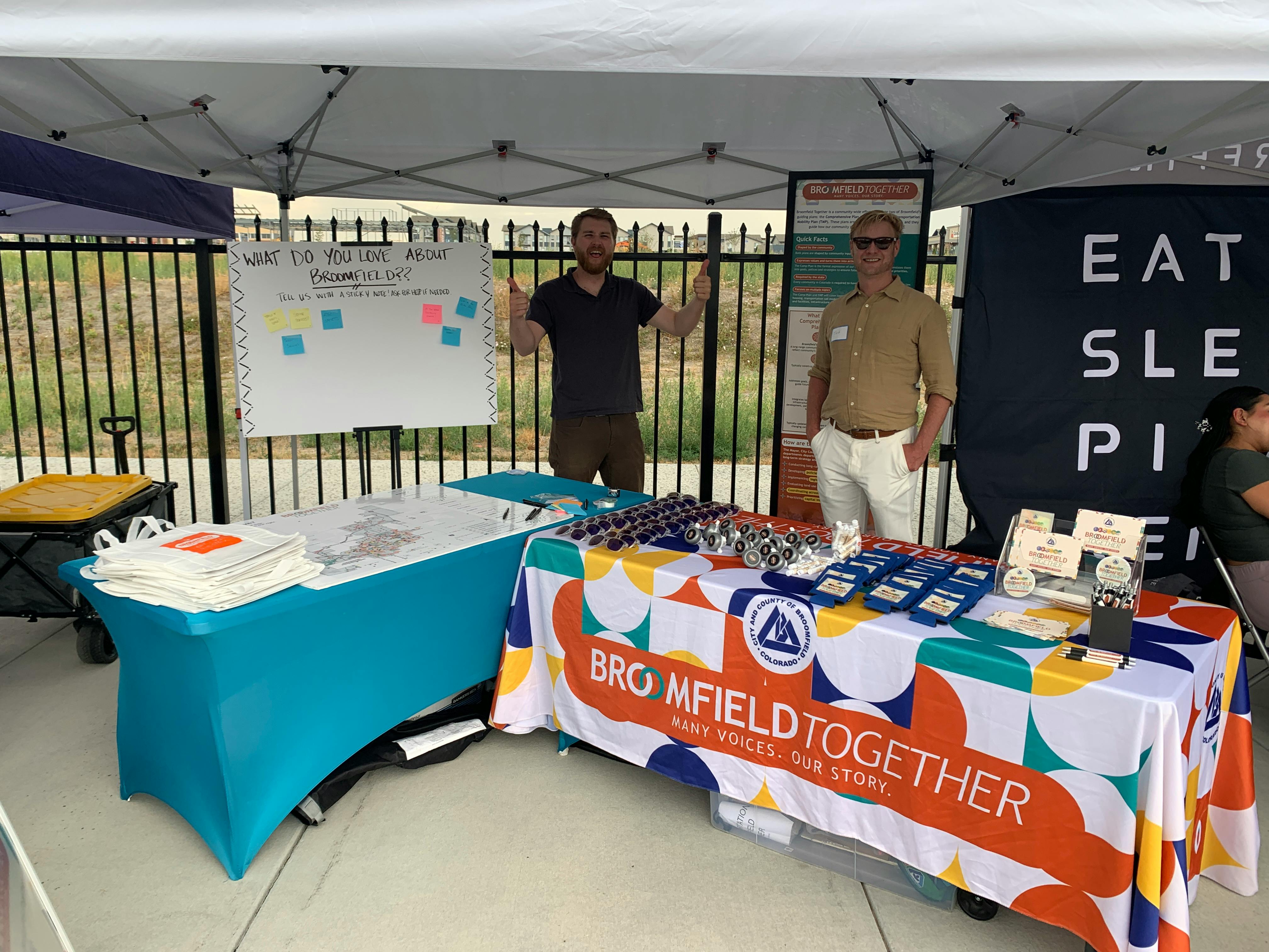 Photograph of two men at a "Broomfield Together" community outreach booth under a white tent. On the left, a man gives two thumbs up next to a whiteboard. On the right, a man in sunglasses stands behind a table covered with a colorful tablecloth.