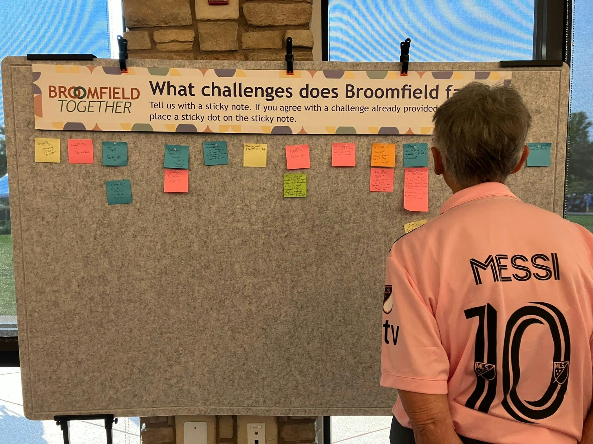 Photograph of a person from behind, wearing a pink Lionel Messi #10 jersey, looking at a community feedback board. A banner at the top of the board reads "BROOMFIELD TOGETHER" and asks, "What challenges does Broomfield face?"