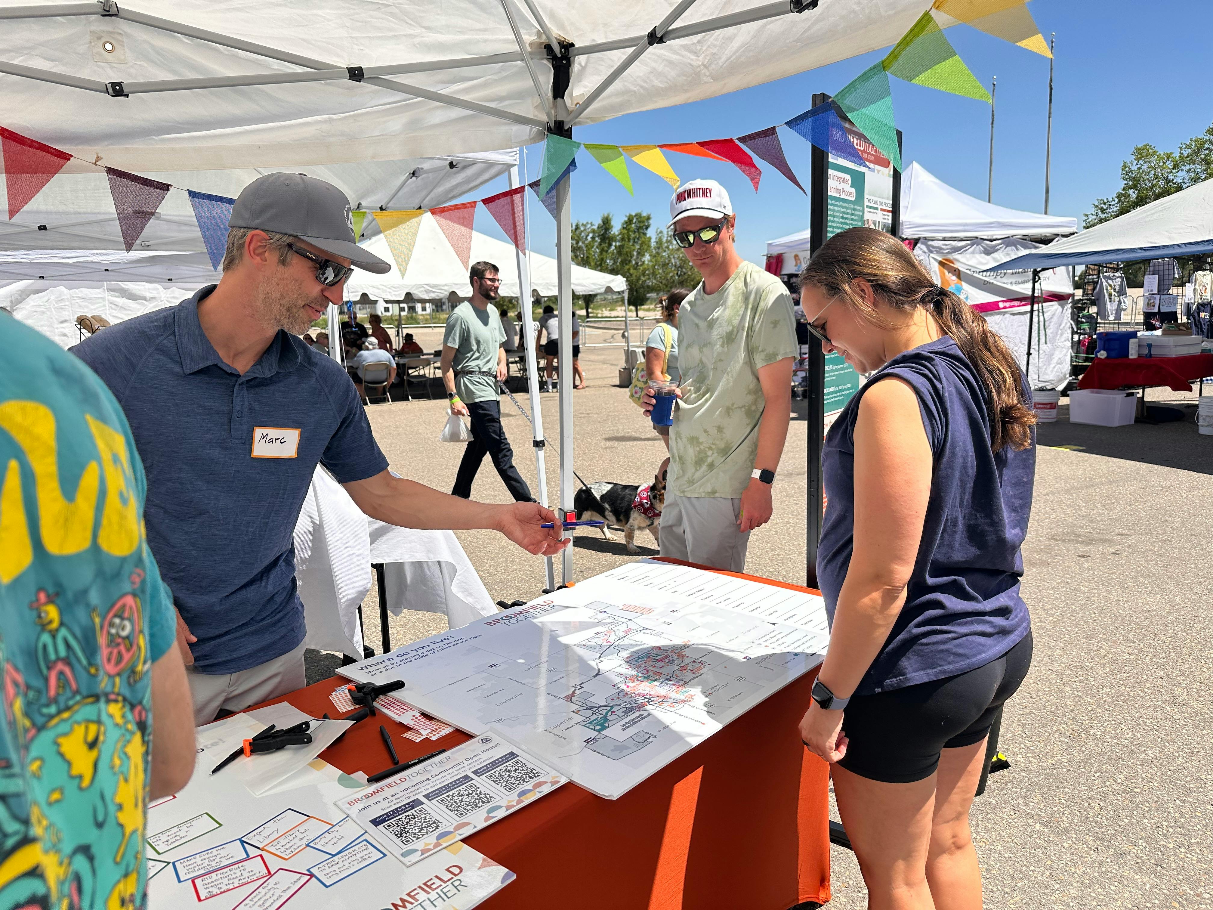 A man, wearing a name tag and a grey cap, points to a large map titled "Broomfield Together" on a table. A woman looks at the map, which asks "Where do you live?"