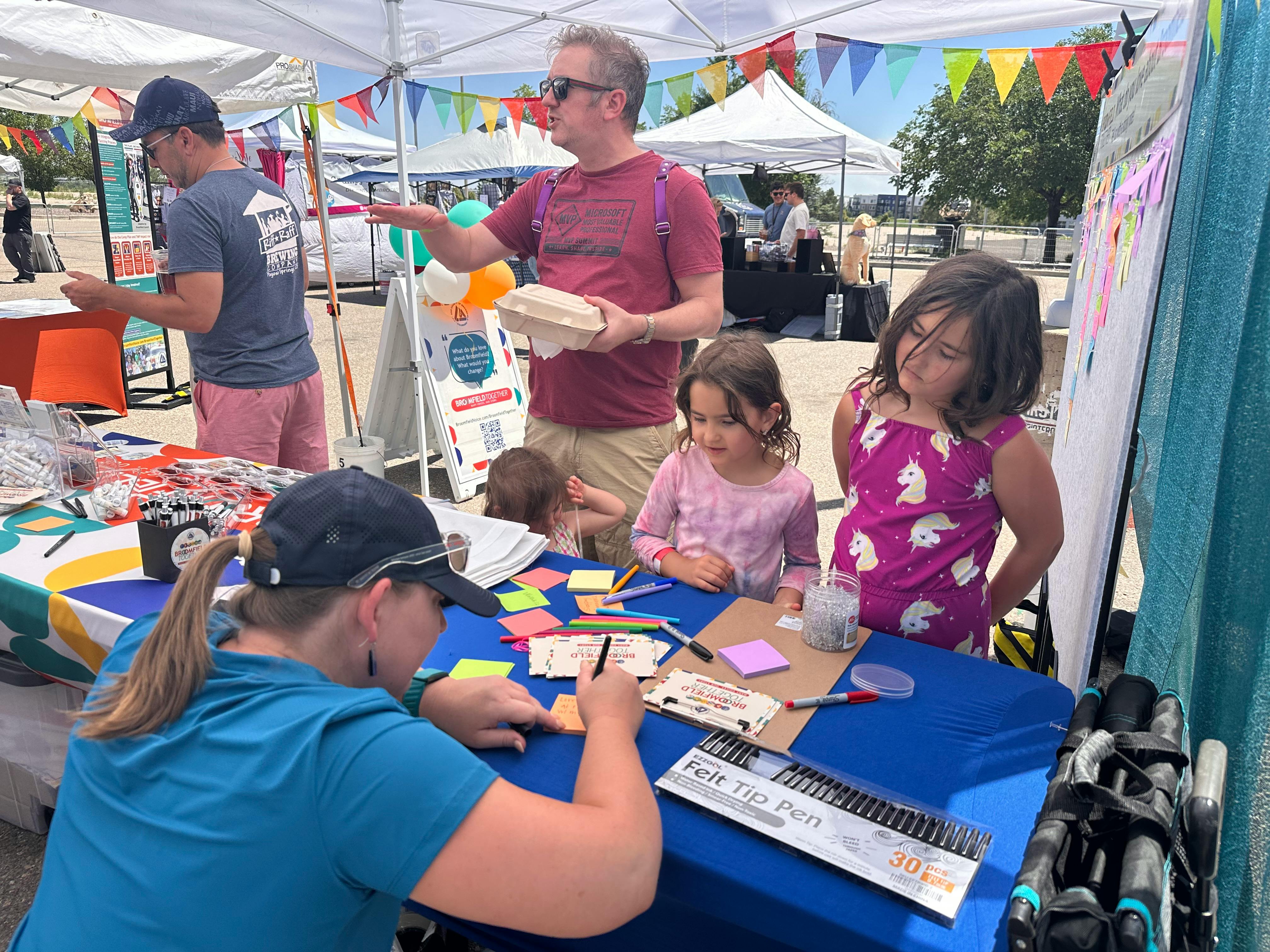 A woman in a blue shirt and baseball cap writes on a card. Two young girls and a man in a red shirt stand behind the table, watching her. The table is covered with a blue cloth and features colorful sticky notes, pens and cards.
