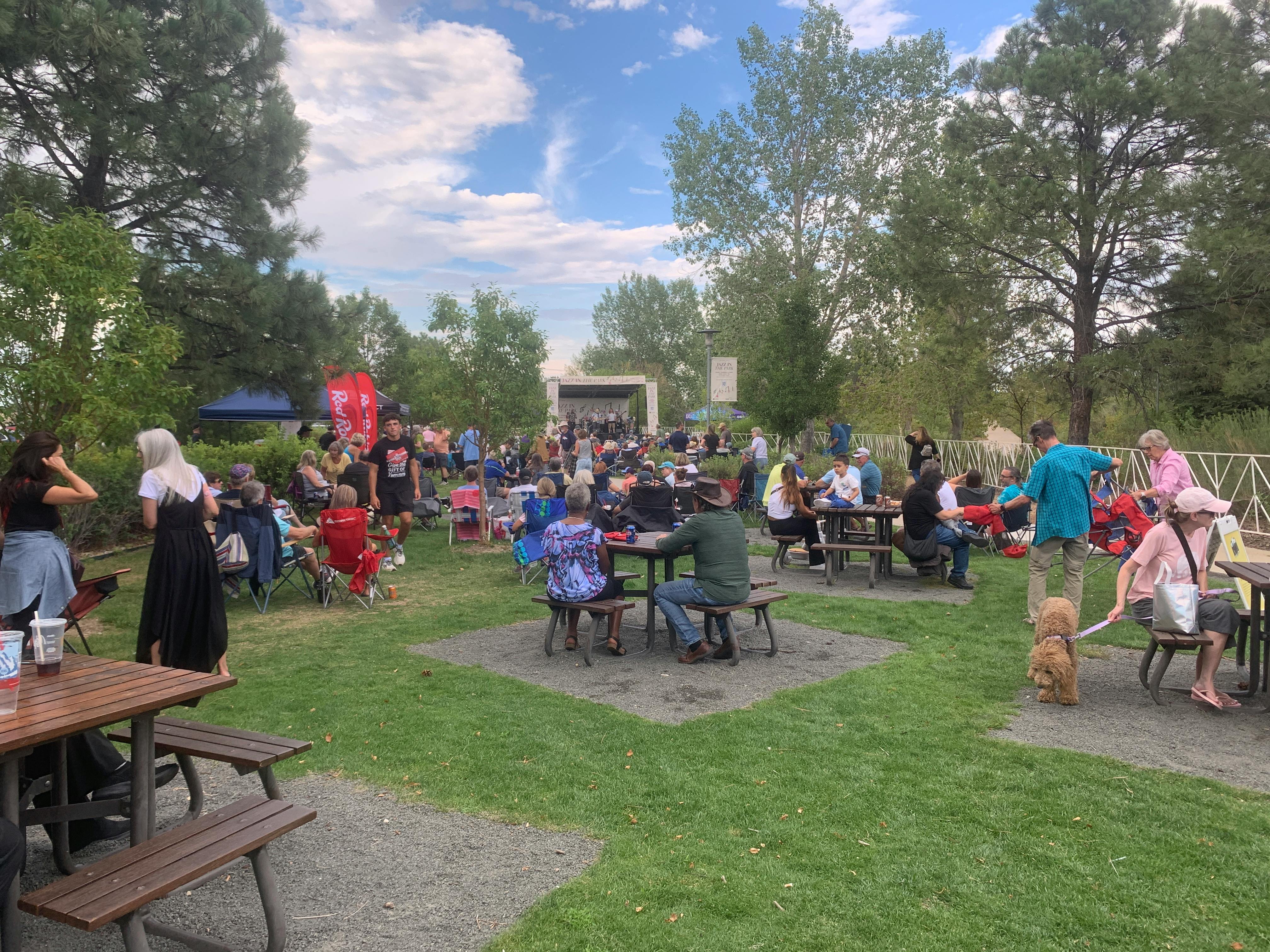 A large crowd of people is gathered on a grassy lawn surrounded by tall trees, sitting at wooden picnic tables and in folding lawn chairs. In the background, a band performs on a white stage under a blue sky with scattered clouds.
